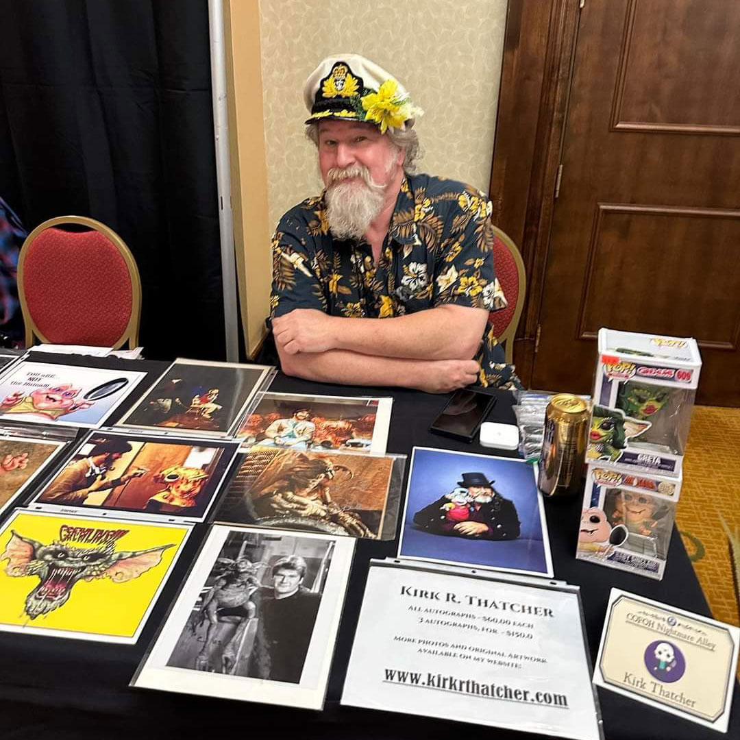 A msiling Kirk Thatcher in a blue and yellow hawaiian shirt shirt wearing a Captains hat and a matching yellow flower in his ear, sitting at his merch table