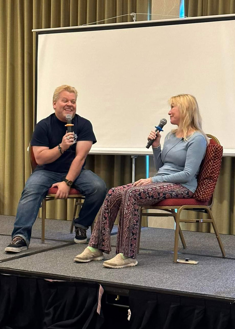 A cute blonde guy, Smurf, in blue jeans, a blue Ghostbuster shirt on, smiling while interviewing a beautiful blonde scream queen in a light blue top and pasiley pants in front of a small projector screen
