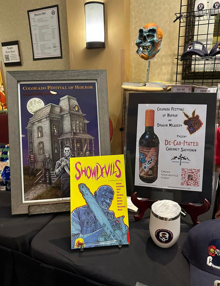 Another side of the merchansizing table with a white wine tumbler, the ShowDevils comic, a print of the Colorado Festival of Horror print for 2023 with slasher killers in front of the Bates house and a special mask made by Daniel Croiser in the background