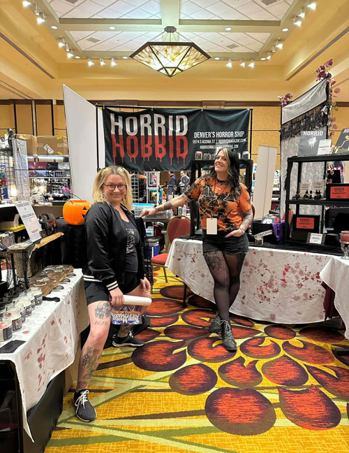 Two ladies vendoing at their booth Horrid, one blonde in glasses smiling with black shorts with her legs stretched out and another tall black ahir with blonde highlights leaning on a table in a black and orange pumpkin tshirt, black shorts and black tights