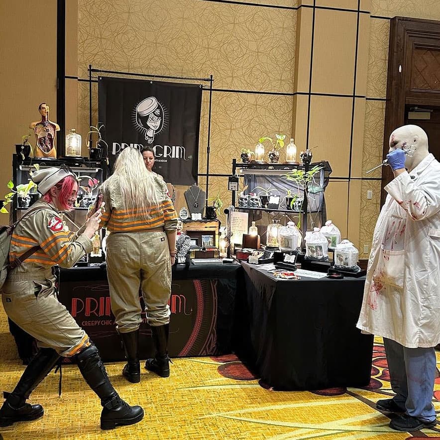 A Ghostbuster with pink hair, in a beigh flight suit with warning tape all over taking a picture of a silent killer doctor who is bald in a messy white coat and blue scrub pants, while another female blonde Ghostbuster in the same outfit is looking at a vendor booth full of plants