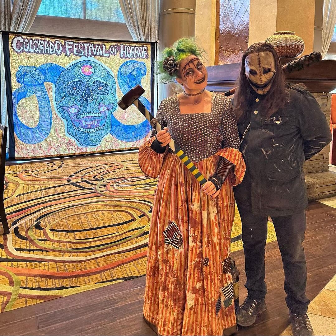 Two guests dressed in killer outfits, one smiling green haired female with a bloody foreheadin a bloody black top polka dot dress with orange stripped bottom, holding a hammer, while a person in all black stands next to her in a dirty white mask