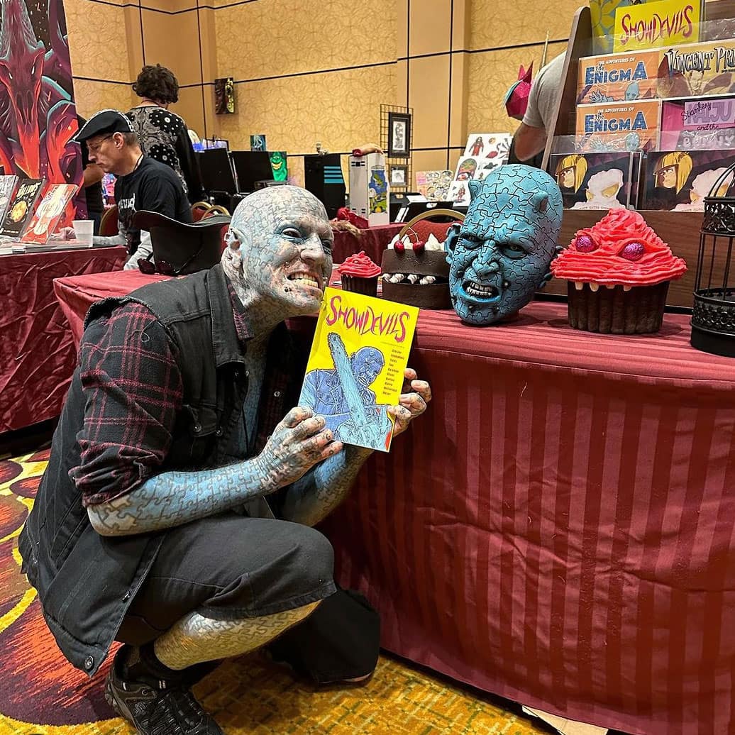 The Enigma, a bald blue man covered in puzzle tattoos and facial piecercings bends down in front of his tabel showing the new ShowDevils comic book with his face on it, he is wearing black shorts, a black vbest and logn sleeve plaid shirt