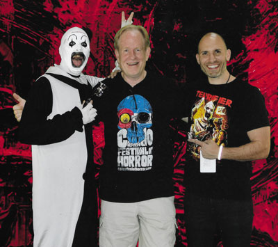 Bret, tall blonde man in a black Colorado Festival of Horror tshirt with skull in white shorts, inbetween Art the Clown, a male clown in black and a man in a black Terrifier shirt in front of a red and black background