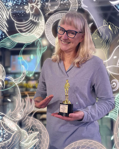 Jeanni, smiling blonde woman with shoulder length hair and black glasses, in a light longsleeve purple shirt, holding an award for Colorado Festival of Horror with an animal filter around the edges